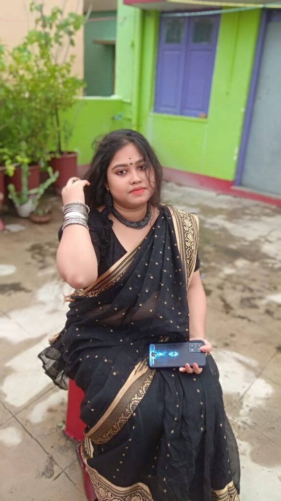 A woman dressed in a black saree with gold embroidery sits on a red stool in an outdoor courtyard. She is accessorized with silver bangles, a black choker necklace, and earrings. Her hair is slightly tousled, and she holds a smartphone in her hand, with a calm expression on her face. The background features potted plants and a colorful green and purple house wall.