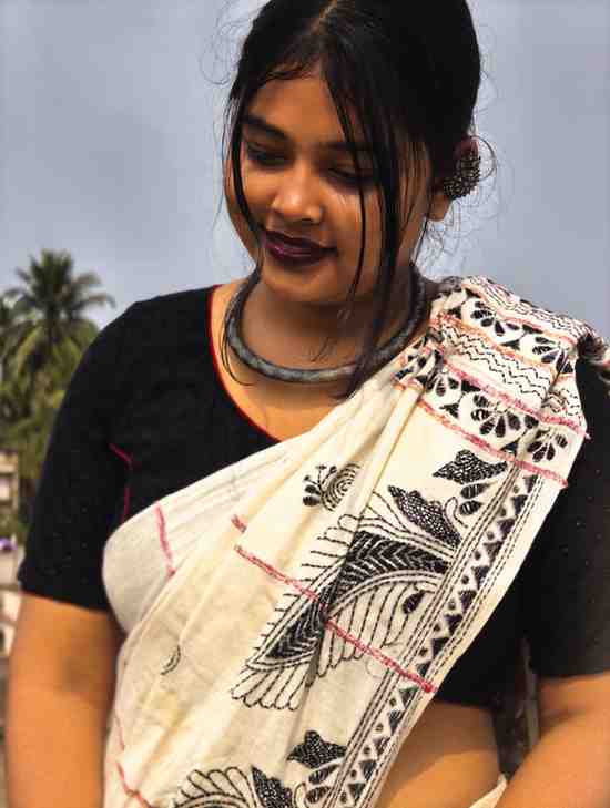 A young woman wearing a traditional white saree with black patterns and a black blouse stands outdoors, looking down with a gentle smile. She has dark lipstick, a simple necklace, and her hair is tied back. Palm trees and a clear blue sky .