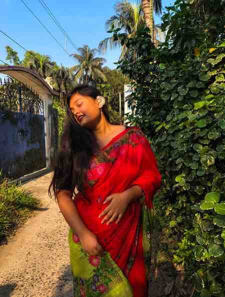 A woman stands outdoors on a sunny day, dressed in a vibrant red and green floral saree. She has long, flowing hair and is wearing a large decorative earring on one ear. Her eyes are closed, and she has a serene expression on her face. The background features lush green foliage, palm trees, a blue gate, and a clear blue sky, creating a peaceful, tropical setting.