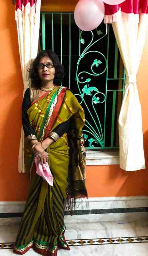 A woman stands indoors in front of a window adorned with white curtains and pink balloons. She is wearing a traditional green and red striped saree with gold and black detailing, paired with a black blouse with gold borders. She accessorizes with gold jewelry, including a necklace, bangles, rings, and a red bindi on her forehead. The background features an orange wall and a decorative green window grill, adding to the festive ambiance. She is holding a pink cloth in her hands.