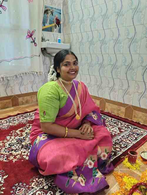 A smiling woman dressed in a vibrant pink and purple saree with floral patterns sits cross-legged on a decorative carpet inside a tiled room. She wears traditional jewelry, including bangles and a necklace, with jasmine flowers adorning her hair. Flower petals and a lit lamp are arranged in front of her, suggesting a festive or ceremonial setting.