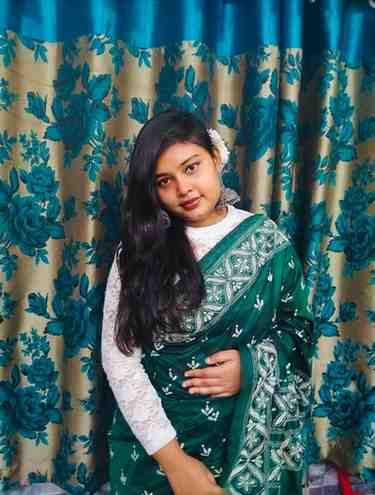 A young woman wearing a green saree with white embroidery and a white lace blouse poses indoors in front of floral-patterned curtains that match the colors of her outfit. She has long, dark hair adorned with a white flower and is wearing large, ornate earrings. She has a gentle smile and a calm expression.