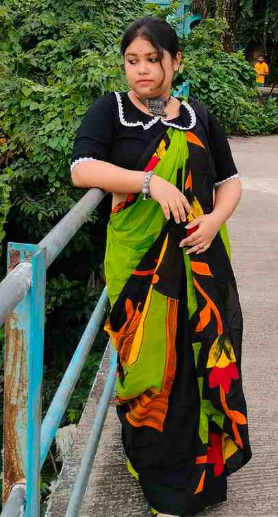 Woman wearing a vibrant black and green saree with bold multicolor prints, standing on a bridge surrounded by greenery.