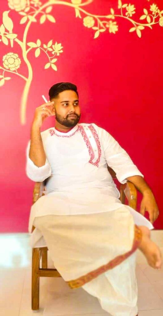 Man in traditional white kurta and dhoti with red embroidery detailing, sitting on a wooden chair against a vibrant red floral wall