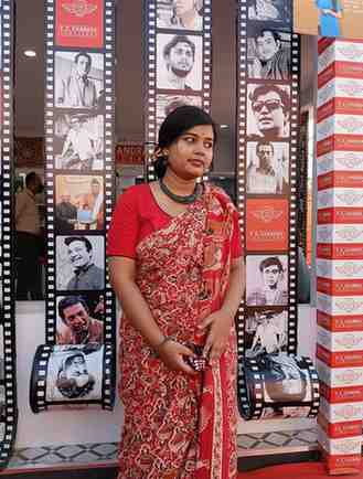 Woman in a traditional red printed saree with black choker necklace, standing in front of a film-themed backdrop featuring classic Indian cinema icons.