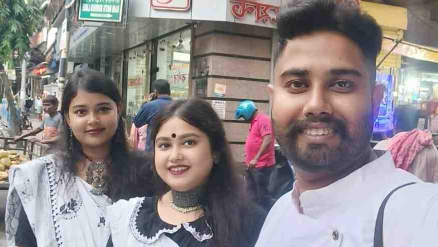 Three people, two women and one man, pose for a selfie on a busy street. The women are dressed in traditional white and black attire with matching jewelry, and the man is wearing a white kurta. They are smiling and appear to be enjoying their time together. The background features shops, signage in Bengali, and street vendors, indicating a lively urban setting.