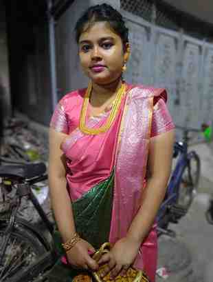 Young woman dressed in a traditional pink and green silk saree with gold jewelry, standing outdoors at night beside a bicycle