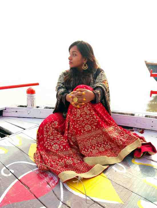 A woman dressed in a richly embroidered red and gold lehenga with a black dupatta sits cross-legged on a colorful wooden boat. She is looking to the side, wearing large golden earrings and a statement ring. The bright sunlight reflects off the water in the background, giving the scene a serene and dreamy ambiance