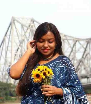 Woman in a blue printed saree holding a bouquet of sunflowers near Howrah Bridge in Kolkata, smiling with eyes closed.