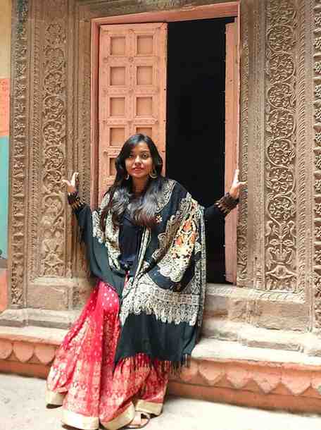 Woman in a traditional red lehenga and embroidered black shawl posing in front of an ornate carved doorway at a heritage site.