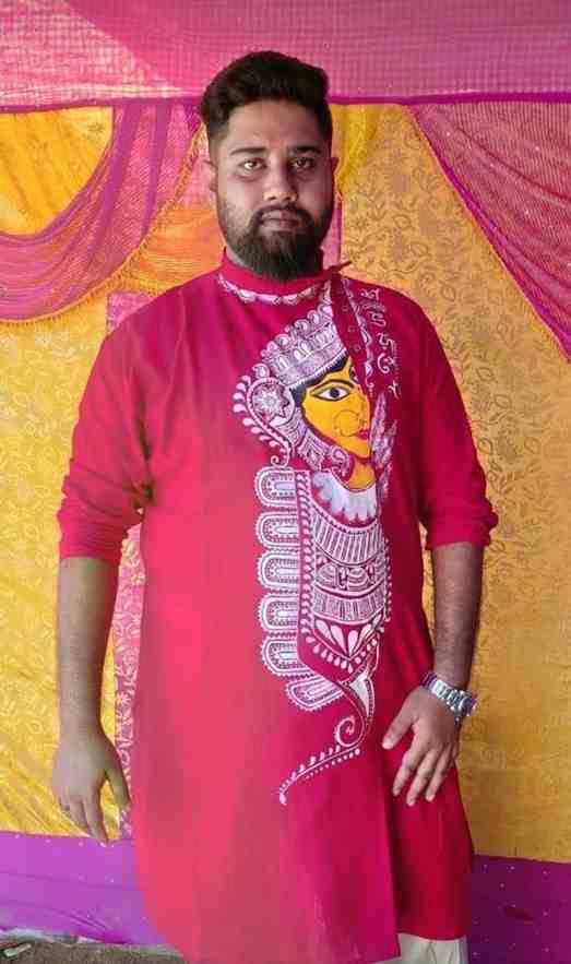 A man wearing a vibrant red kurta with an artistic traditional face design stands in front of a colorful backdrop featuring yellow, pink, and purple fabric drapes. He has a neatly groomed beard, short hair, and is wearing a silver wristwatch, looking calmly at the camera.