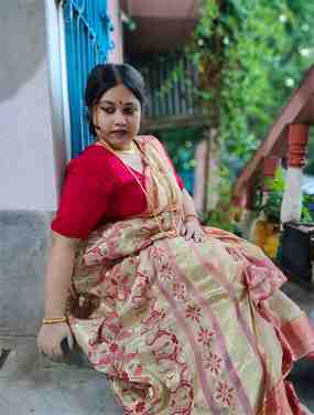 A woman dressed in a traditional cream and red saree with gold and floral patterns sits gracefully near a blue gate and staircase. She is adorned with gold jewelry, including necklaces and bangles, and has a red bindi on her forehead. The background is a mix of greenery and a residential setting, creating a serene and cultural ambiance.