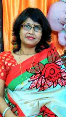 Woman wearing a white saree with red and green floral patterns and a red blouse, seated indoors in front of an orange curtain with a soft toy in the background.