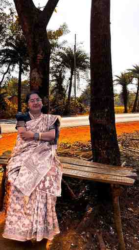 An elderly woman wearing a traditional saree sits on a rustic wooden bench under tall trees near a roadside. She is smiling, arms crossed, and appears relaxed. The background features a rural landscape with red soil, palm trees, and a sunlit road.