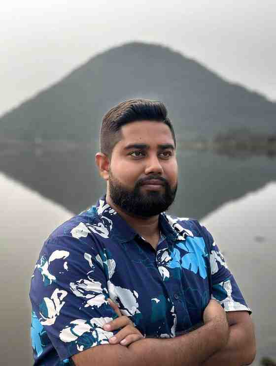A man standing with arms crossed in front of a calm lake, with a mountain and its reflection in the water forming a symmetrical backdrop. He is wearing a navy blue floral shirt and has a well-groomed beard and short hair, looking thoughtfully into the distance.
