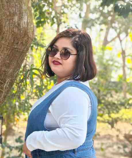 A woman standing outdoors in a sunlight, green environment, posing beside a tree. She is wearing a white long-sleeve top under a denim overall, accessorized with round sunglasses and hoop earrings. Her short, dark hair is styled neatly, and she has a confident expression with a touch of red lipstick.