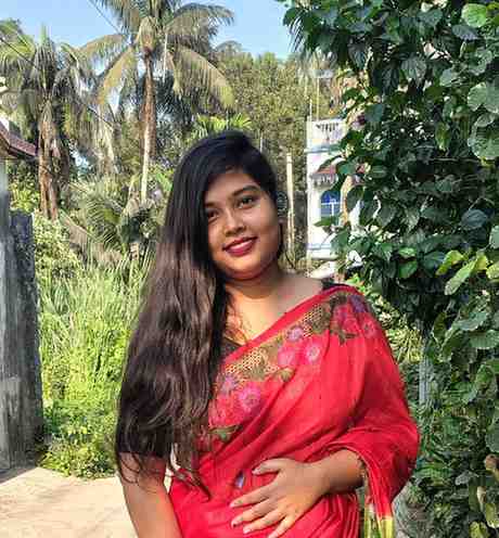 A woman standing outdoors on a sunny day, surrounded by lush greenery and palm trees. She is wearing a bright red saree adorned with floral embroidery and has long, flowing black hair. She is smiling warmly at the camera, creating a cheerful and vibrant atmosphere.