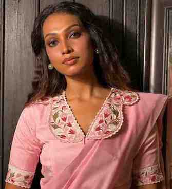 Woman wearing a pink designer saree with floral embroidered blouse, posing against a dark wooden background.
