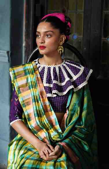 A woman wearing a colorful traditional Indian saree with a checked pattern in green, yellow, and pink hues, paired with a purple blouse featuring a distinctive white lace collar. She is seated indoors, accessorized with gold earrings and a pink hairband, looking poised and elegant.