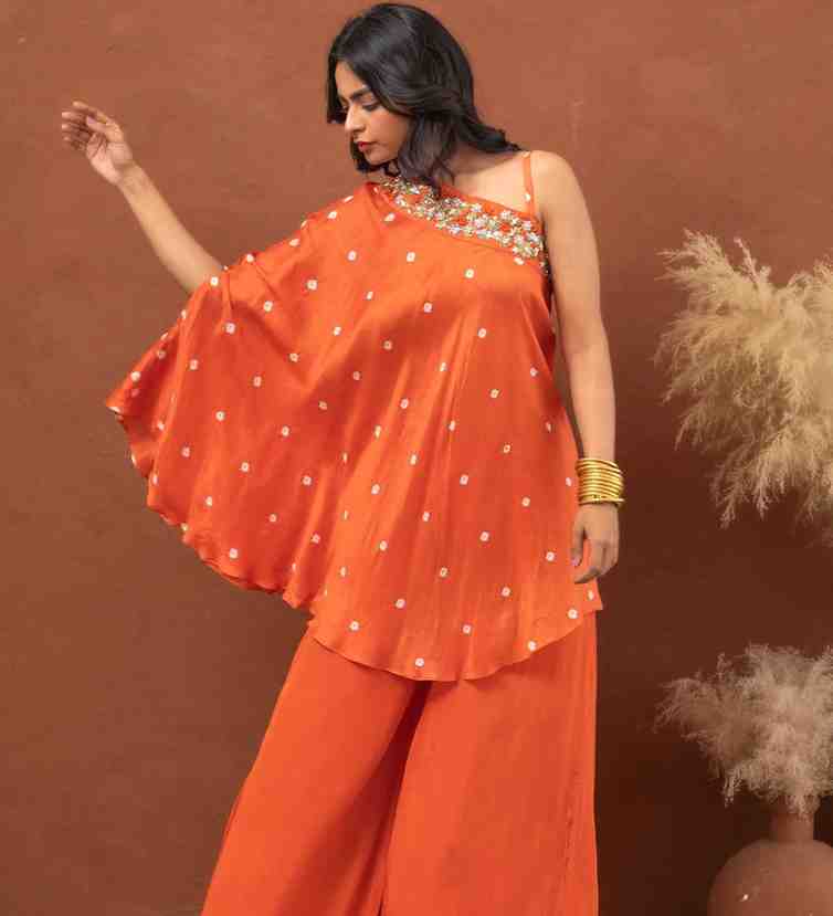 Woman wearing an orange asymmetrical one-shoulder co-ord set with floral embroidery and wide-leg pants, styled with gold bangles.