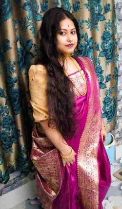 A woman dressed in a traditional Indian saree poses indoors. She is wearing a rich magenta and gold silk saree with intricate patterns, paired with a beige blouse. Her long, dark, wavy hair is left open, and she accessorizes with gold jewelry including bangles, a necklace, earrings, and a nose ring. She stands in front of a floral curtain and patterned tiled floor, exuding elegance and cultural grace.