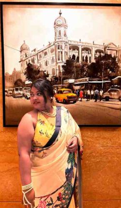 A woman poses gracefully indoors in front of a large framed painting depicting an old colonial building with a clock tower and vintage vehicles on the street. She is wearing a cream-colored saree adorned with floral and peacock motifs, paired with a sleeveless yellow floral blouse. Her hair is tied back in a loose bun, and she accessorizes with pearl jewelry including a necklace and bracelets. The background features warm-toned marble walls, adding a touch of elegance to the setting.